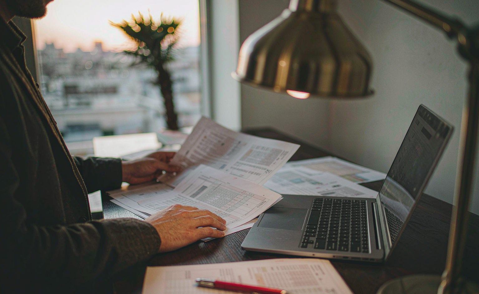 Hands reviewing printed trade documents beside a laptop and a pen on a desk in warm office light.